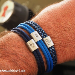 Könnte beinhalten: Drei geflochtene Armbänder aus Seil mit silbernen Metallverschlüssen. Jeder Verschluss hat ein anderes Symbol eingraviert. Die Armbänder sind blau und schwarz.