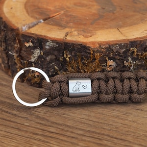 Könnte beinhalten: Brauner Paracord-Schlüsselanhänger mit silberfarbenem Metallakzent. Das Metallstück zeigt eine Hundesilhouette und ein Herzsymbol. Der Schlüsselanhänger hat einen silberfarbenen Schlüsselring zum Befestigen von Schlüsseln.