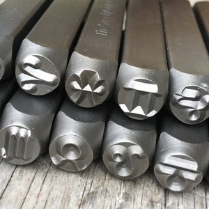 Könnte beinhalten: Ein Satz von 12 Metallstempeln mit verschiedenen Tierkreiszeichen. Die Stempel sind in einem Stapel angeordnet, wobei die Symbole nach oben zeigen. Die Symbole sind alle kreisförmig und aus dunkelgrauem Metall.