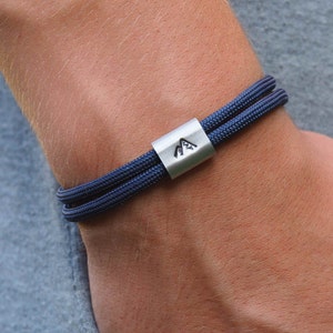 Könnte beinhalten: Ein dunkelblaues Paracord-Armband mit einem silbernen Metallverschluss, der ein Berglogo trägt.
