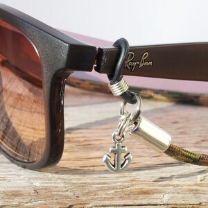 Op de afbeelding: Een close-up van een paar bruine Ray-Ban zonnebrillen met een camouflagekoord en een ankerbedel eraan bevestigd. De zonnebril ligt op een houten oppervlak.