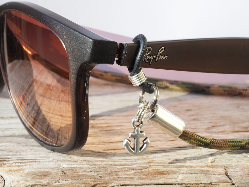 Op de afbeelding: Een close-up van een paar bruine Ray-Ban zonnebrillen met een camouflagekoord en een ankerbedel eraan bevestigd. De zonnebril ligt op een houten oppervlak.