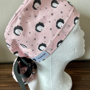 May include: Pink surgical scrub cap with a black and white print of a woman's face and stars. The cap has a black tie and a white label that says "Willowbird Caps".