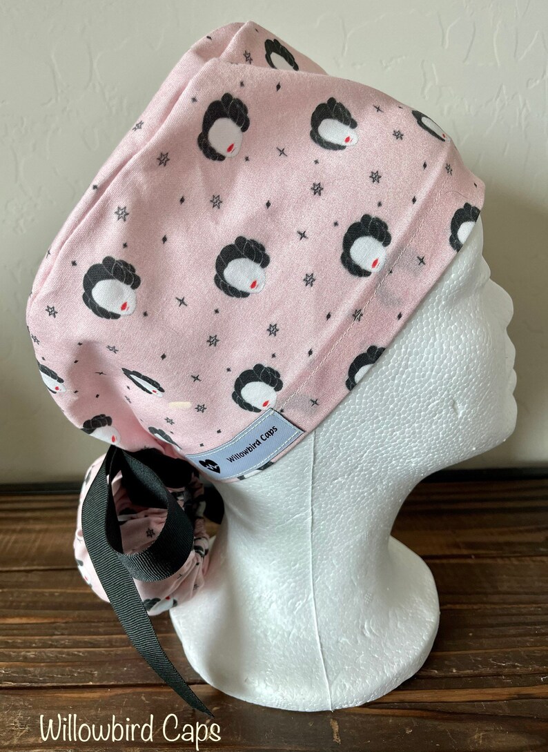 May include: Pink surgical scrub cap with a black and white print of a woman's face and stars. The cap has a black tie and a white label that says "Willowbird Caps".