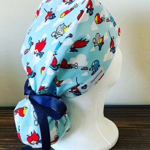 May include: A blue and white scrub hat with a tie at the back. The hat has a pattern of cartoon superheroes flying in the sky. The text "Willowbird Caps" is on the hat.