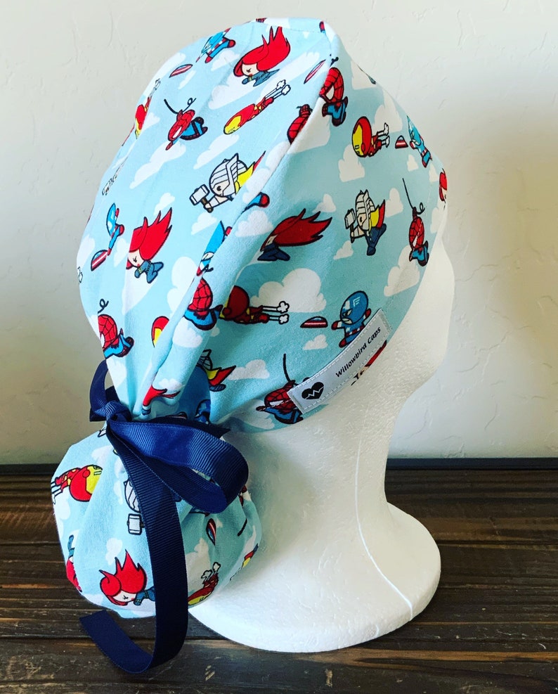 May include: A blue and white scrub hat with a tie at the back. The hat has a pattern of cartoon superheroes flying in the sky. The text "Willowbird Caps" is on the hat.