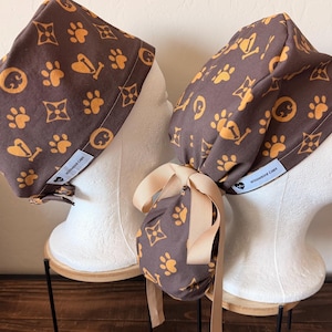 Puede incluir: Dos gorros quirúrgicos marrones con un patrón naranja repetitivo de huellas, huesos y estrellas de cuatro puntas. Cada gorro tiene una etiqueta de tela blanca con el texto Willowbird Caps y un pequeño icono de corazón. Un gorro incluye un lazo beige.