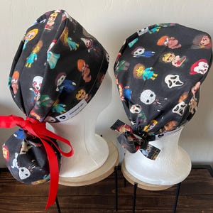 May include: Two black scrub hats with a colorful print of horror movie characters. The hats are tied with red and black ribbons. The hats are on white mannequins with wooden bases.