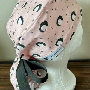 May include: Pink surgical scrub hat with a black and white print of a woman's face. The hat has a black ribbon tie and a white label that says "Willowbird Caps".