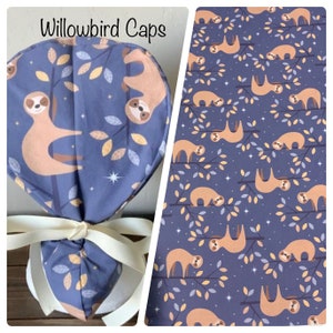 May include: A blue surgical cap with a white tie, featuring a repeating pattern of sloths hanging from branches with yellow leaves and white stars. The text "Willowbird Caps" is printed on the cap.