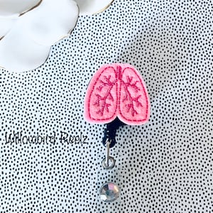 May include: A pink felt lung-shaped badge reel with embroidered details. The badge reel has a black cord, a silver-tone metal clip, and a clear plastic holder with a decorative gem. The background is a black and white polka dot pattern.