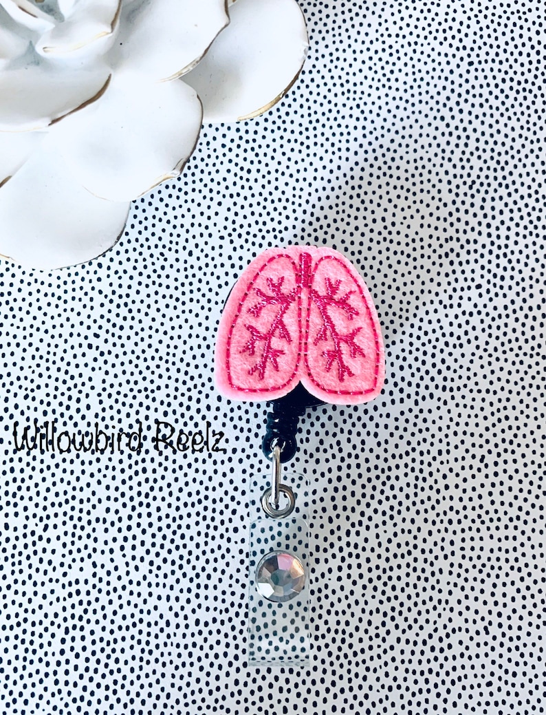 May include: A pink felt lung-shaped badge reel with embroidered details. The badge reel has a black cord, a silver-tone metal clip, and a clear plastic holder with a decorative gem. The background is a black and white polka dot pattern.
