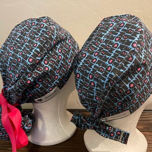 Op de afbeelding: Twee blauwe en zwarte gepatroneerde chirurgische scrubcaps met roze harten en het woord "BE KIND" dat zich herhaalt. Een cap heeft een roze lint dat aan de achterkant is vastgebonden.