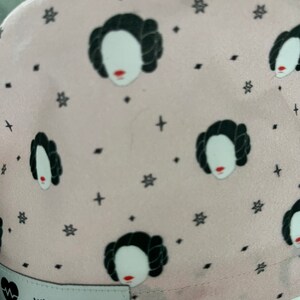 May include: Pink fabric hat with a repeating pattern of a woman's face with a black bun and a white background. The hat has a black label that says "Willowbird Caps".
