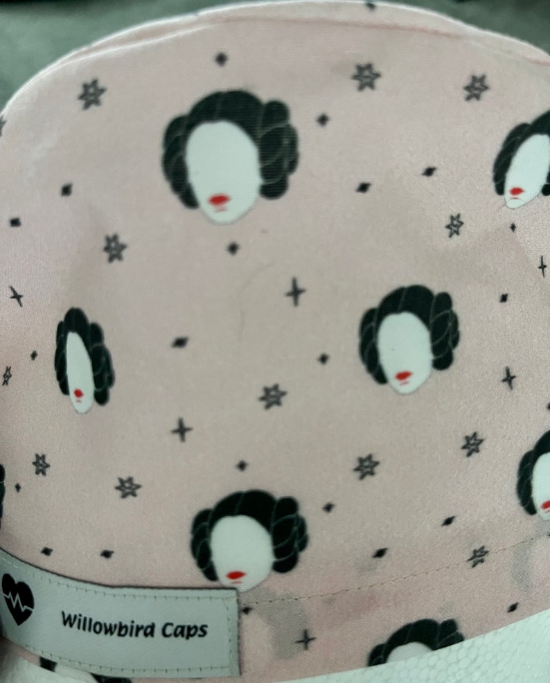 May include: Pink fabric hat with a repeating pattern of a woman's face with a black bun and a white background. The hat has a black label that says "Willowbird Caps".
