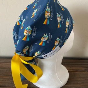 Könnte beinhalten: Eine blaue OP-Mütze mit gelber Schleife. Die Mütze hat ein sich wiederholendes Muster des Raumschiffs Millennium Falcon mit dem Text "Millennium Falcon" auf jedem Raumschiff. Die Mütze hat ein weißes Etikett mit dem Text "Willowbird Caps" und einem Herzsymbol.