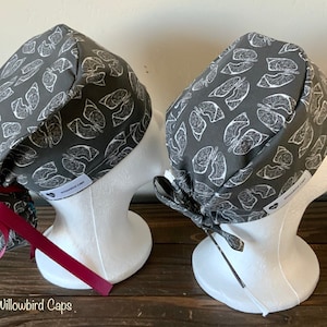 Könnte beinhalten: Zwei graue OP-Hauben mit einem weißen anatomischen Lungen-Druck. Die Hauben haben eine burgunderfarbene Schleife. Die Hauben befinden sich auf weißen Mannequin-Köpfen. Der Text "Willowbird Caps" befindet sich am unteren Rand des Bildes.
