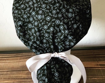 Könnte beinhalten: Eine schwarze OP-Mütze mit weißem Blumenmuster und einer weißen Schleife. Willowbird Caps ist auf der Holzoberfläche unter der Mütze aufgedruckt.