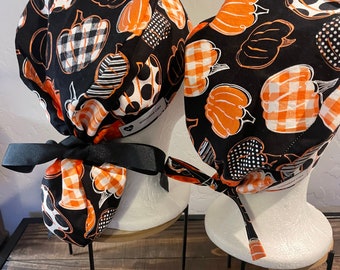 Pode incluir: Dois gorros de cirurgia pretos e laranja com um padrão xadrez branco e laranja e bolinhas pretas. Os gorros são amarrados com uma fita preta e apresentam um design de abóbora.