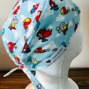 May include: A light blue surgical scrub cap with a white tie. The cap features a repeating pattern of cartoon superheroes on a white cloud background. The text "Willowbird Caps" is printed on the side of the cap.