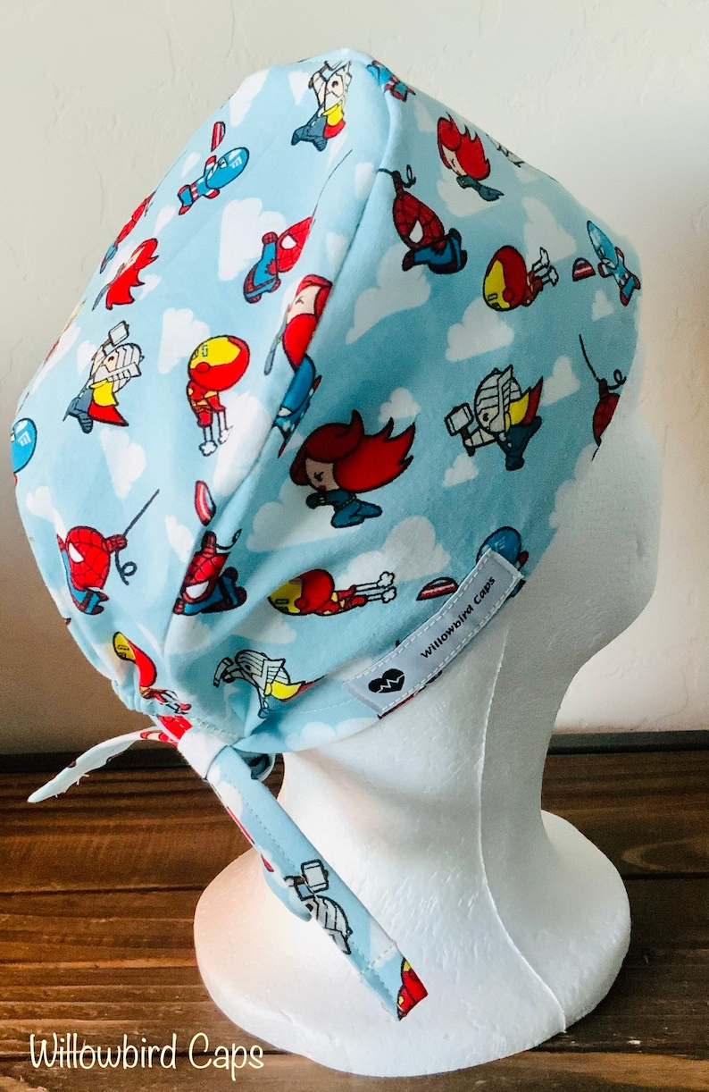 May include: A light blue surgical scrub cap with a white tie. The cap features a repeating pattern of cartoon superheroes on a white cloud background. The text "Willowbird Caps" is printed on the side of the cap.