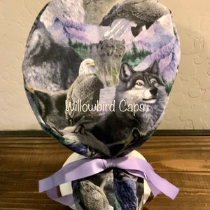 May include: A purple scrub hat with a print of a wolf, eagle, and mountains. The hat has a purple ribbon tied around it. The text "Willowbird Caps" is printed on the hat.