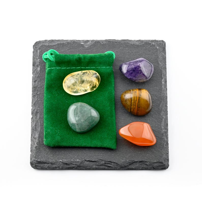 May include: A green velvet drawstring pouch containing a yellow citrine stone and a smooth green stone, accompanied by three loose polished stones in purple, brown tiger eye, and orange, all arranged on a square dark gray slate tile.