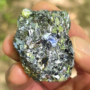 May include: A close-up of a mineral specimen with a metallic luster. The rock has a dark gray base color with iridescent patches of blue, green, and gold. The surface is rough and uneven, with sharp edges and crystalline structures.