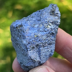 May include: A dark blue mineral specimen with a metallic sheen. The rough, irregular surface reflects light, showing streaks of silver and gold. The rock is held in a hand against a blurred green background.