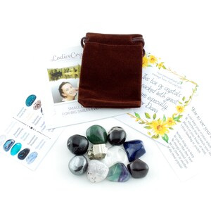May include: A collection of polished gemstones in various colors, including green, black, blue, and clear, alongside a small brown velvet pouch and informational cards. The gemstones are arranged on a white surface, creating a visually appealing display.