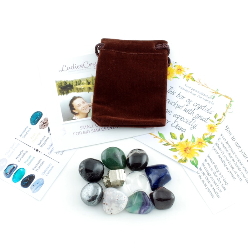 May include: A collection of polished gemstones in various colors, including green, black, blue, and clear, alongside a small brown velvet pouch and informational cards. The gemstones are arranged on a white surface, creating a visually appealing display.