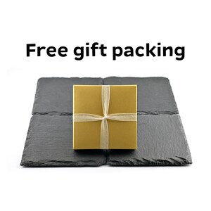May include: A square gold gift box tied with a thin, translucent ribbon, centered on four dark gray, textured slate coasters arranged in a square formation. The text "Free gift packing" appears in black letters above the items.