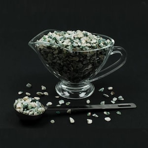May include: A clear glass gravy boat filled with small, green and white speckled stones. A stainless steel measuring spoon holds a portion of the stones. Scattered stones are on the black surface.