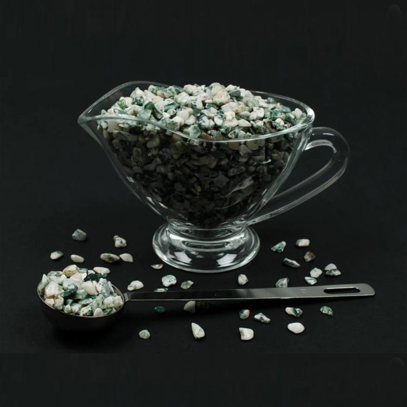 May include: A clear glass gravy boat filled with small, green and white speckled stones. A stainless steel measuring spoon holds a portion of the stones. Scattered stones are on the black surface.