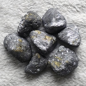 May include: A collection of seven irregularly shaped, dark gray stones with metallic silver and gold accents. The stones are arranged in a circular pattern against a soft, white, textured background. The stones vary in size and have a rough, natural appearance.