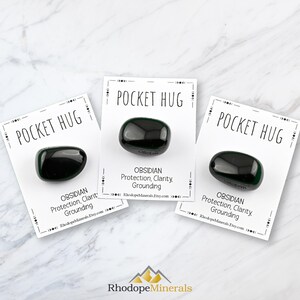 以下が含まれることがあります： 白いカードに「POCKET HUG」と「OBSIDIAN Protection, Clarity, Grounding」のテキストとウェブサイトアドレス「RhodopeMinerals.Etsy.com」が書かれた、黒いオブシディアンのポケットハグストーンが3つあります。