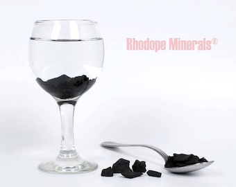 May include: A glass of water with black charcoal pieces in it. A spoon with more charcoal pieces is on a white surface. The text "Rhodope Minerals®" is in the upper right corner of the image.