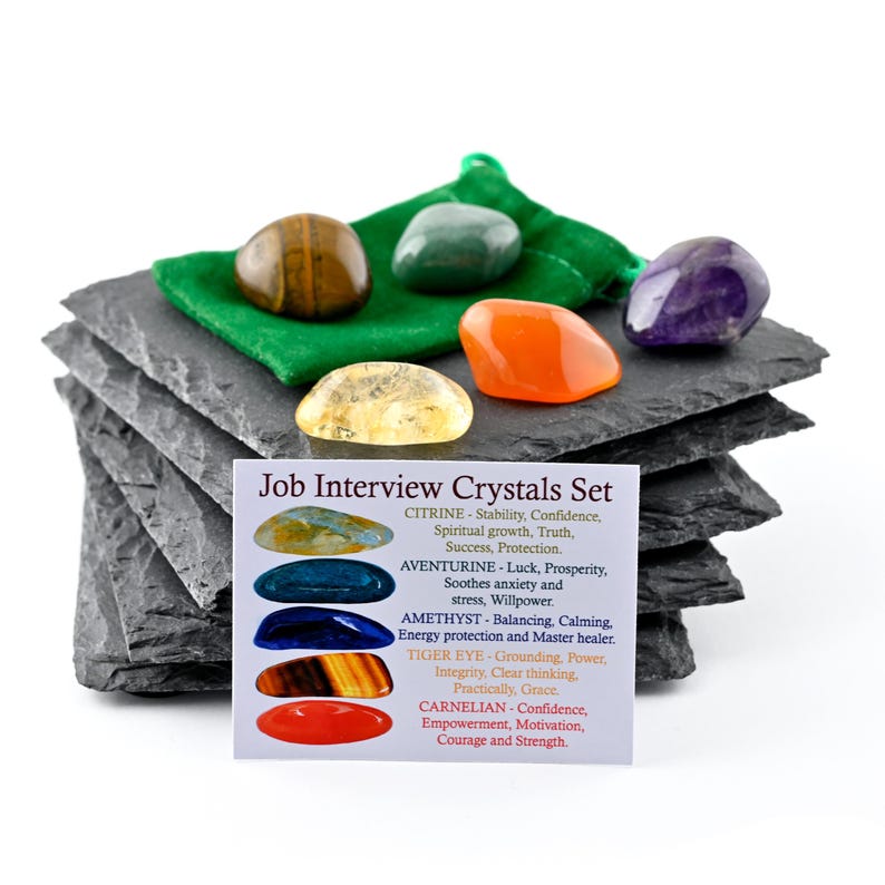 May include: A set of five polished gemstones including tiger eye, aventurine, amethyst, citrine, and carnelian, resting on a stack of dark gray slate tiles with a green fabric pouch and an informational card titled Job Interview Crystals Set.