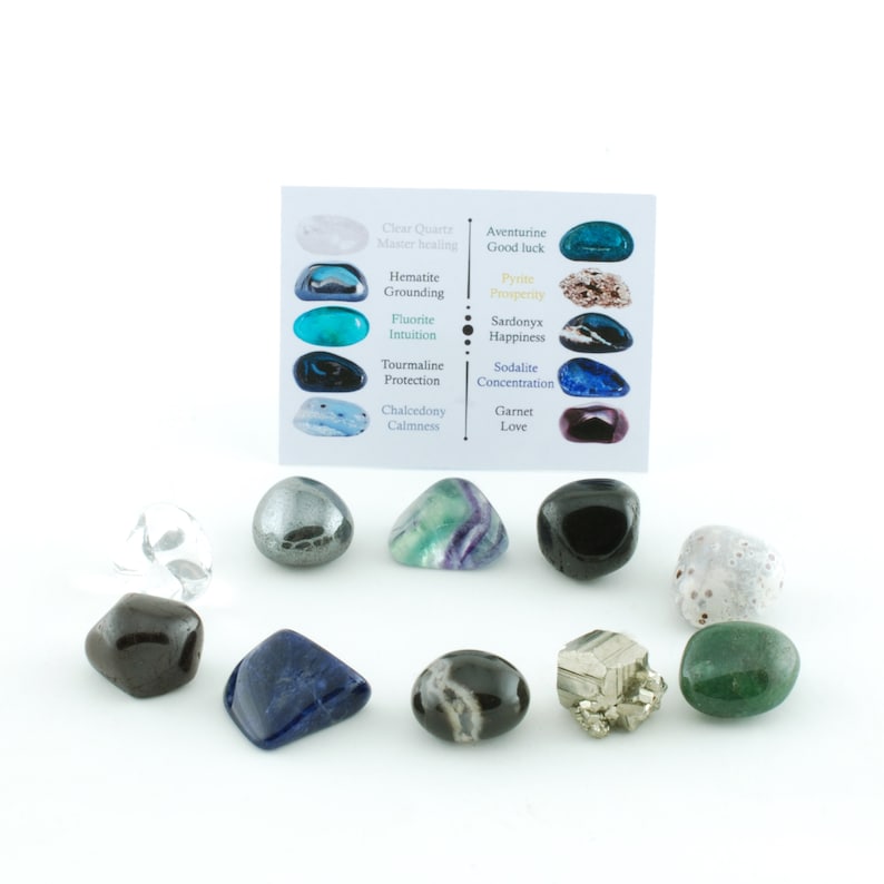 May include: A collection of polished gemstones in various colors, including clear quartz, hematite, and fluorite, arranged on a white surface. A small card lists the names and properties of each stone, such as Aventurine for good luck and Garnet for love.