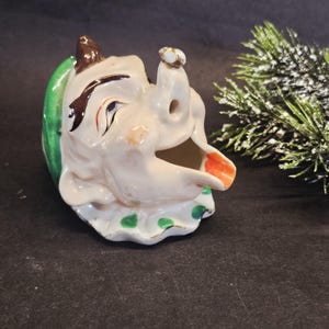 May include: A vintage ceramic clown head creamer with a whimsical design. The clown features a white face, brown hair, and a green collar with green polka dots. The clown's mouth is open, revealing an orange interior.
