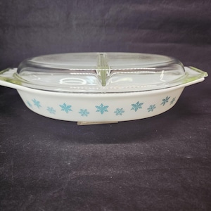 May include: White oval casserole dish with a clear glass lid. The dish features a turquoise snowflake pattern around the rim and has green accents on the handles. The lid is divided into two sections.