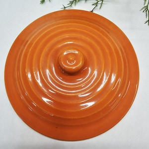 May include: A round, bright orange ceramic lid featuring a glossy finish and a series of concentric circular ridges radiating from a central, raised knob handle. The lid has a smooth, uniform surface texture and a vibrant, solid orange color.