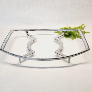 Puede incluir: Un soporte cromado para platos de servir con un marco rectangular y soportes curvos. El soporte tiene un diseño decorativo y ondulado en el centro y se eleva mediante cuatro patas cortas. Una ramita de helecho verde está en el fondo.