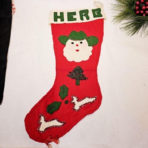May include: Red felt Christmas stocking with the name "HERB" in green letters. The stocking features a Santa Claus face, a beaded Christmas tree, and other festive embellishments. A loop is attached for hanging.