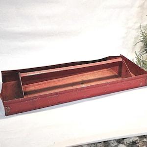 May include: A vintage red metal tool tray with a handle and dividers. The rectangular tray shows signs of wear, with a slightly distressed appearance. The handle is centered, and the dividers create separate compartments.