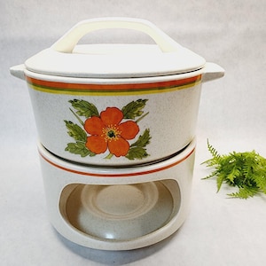 Op de afbeelding: Crèmekleurige keramische fonduepan met deksel en handvat. De pan heeft een bloemenontwerp met een oranje bloem en groene bladeren. Er zijn oranje en gele strepen rond de bovenkant. De pan staat op een bijpassende basis met een uitsparing voor een warmtebron.