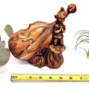 Könnte beinhalten: Ein brauner Keramik-Blumentopf in Geigenform mit einer geschnitzten Elfenfigur. Der Elf kniet mit gefalteten Händen. Der Pflanztopf ist etwa 15 cm lang.