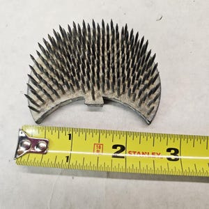 May include: A silver metal flower frog with numerous sharp, black pins. The frog is crescent-shaped and measures approximately 3 inches across. A yellow tape measure is placed next to the frog for scale.