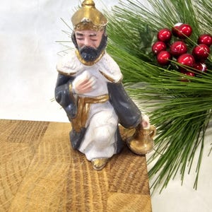 May include: A kneeling figurine of a man with a beard, wearing a gold crown, white robe, and black and gold accents. The figurine is holding a gold vessel. Christmas decor with red berries and green pine branches are in the background.