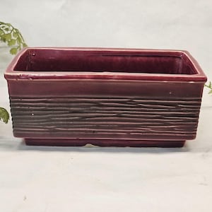 Puede incluir: Una jardinera rectangular de cerámica burdeos con un diseño texturizado similar a la veta de la madera en la parte delantera. La jardinera tiene un borde liso y una base ligeramente elevada. Ideal para suculentas o plantas pequeñas.